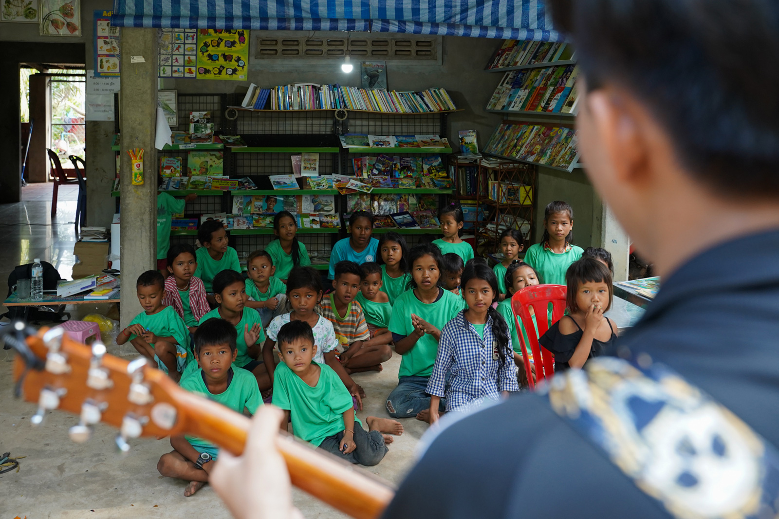 A youth volunteer from Siem Reap City plays guitar for students at the Village Library in Areak Svay Village after learning about the project on social media.